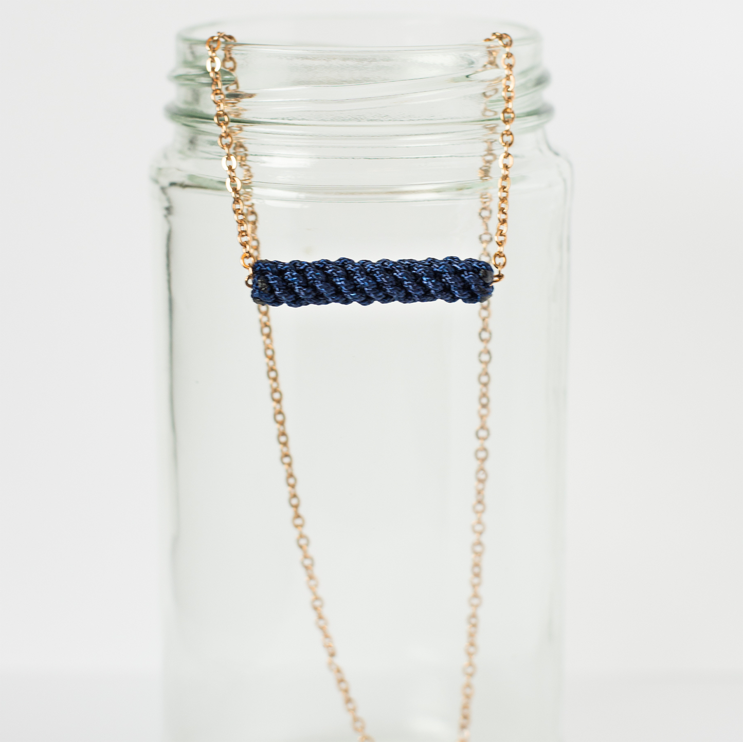 Gold chain necklace with navy braided bar pendant displayed on clear glass jar