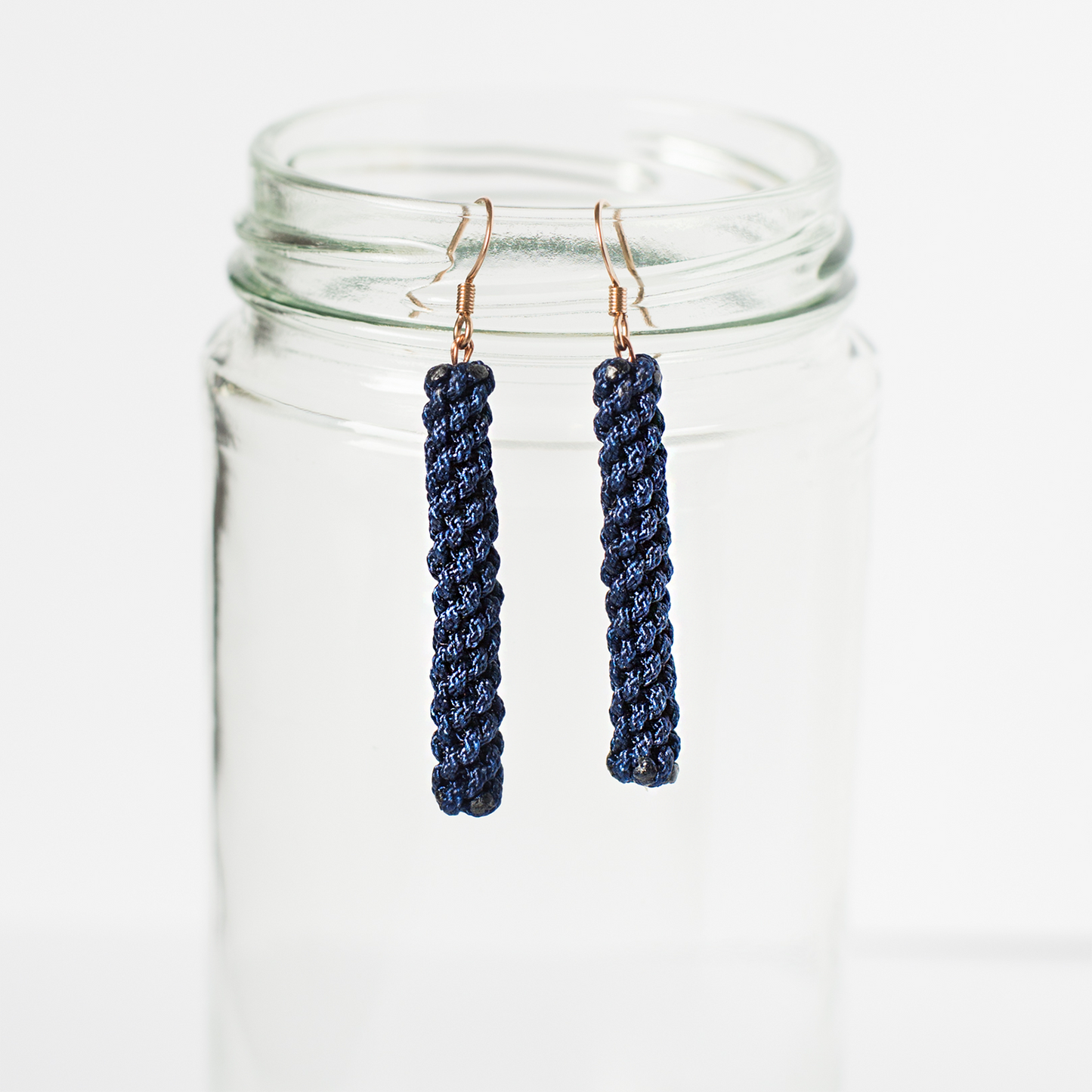 Handmade navy blue woven earrings with gold hooks displayed on a clear glass jar